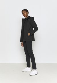 Black waterproof jacket with a hood, zip closure, and side pockets, paired with black trousers and white athletic shoes.