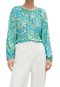 Blouse bleue à motifs floraux verts et blancs, décolleté froncé et manches longues, assortie à un pantalon taille haute blanc.