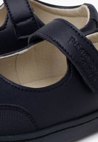 Zapatos Mary Jane de piel negra con una correa de velcro, punta redonda y un interior acolchado. Presentan detalles texturizados y costuras decorativas.
