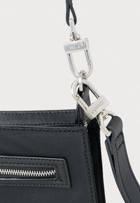 Μαύρη νάιλον τσάντα ώμου με λεία υφή, ασημένια μεταλλικά στοιχεία, τσέπη με φερμουάρ και ρυθμιζόμενο ιμάντα που στερεώνεται με κλιπ καραμπίνερ.