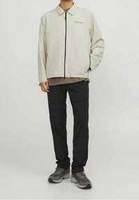 Giacca zip-up beige chiaro con colletto, pantaloni cargo neri e sneakers grigie. La giacca presenta una stampa del logo e tasche laterali.