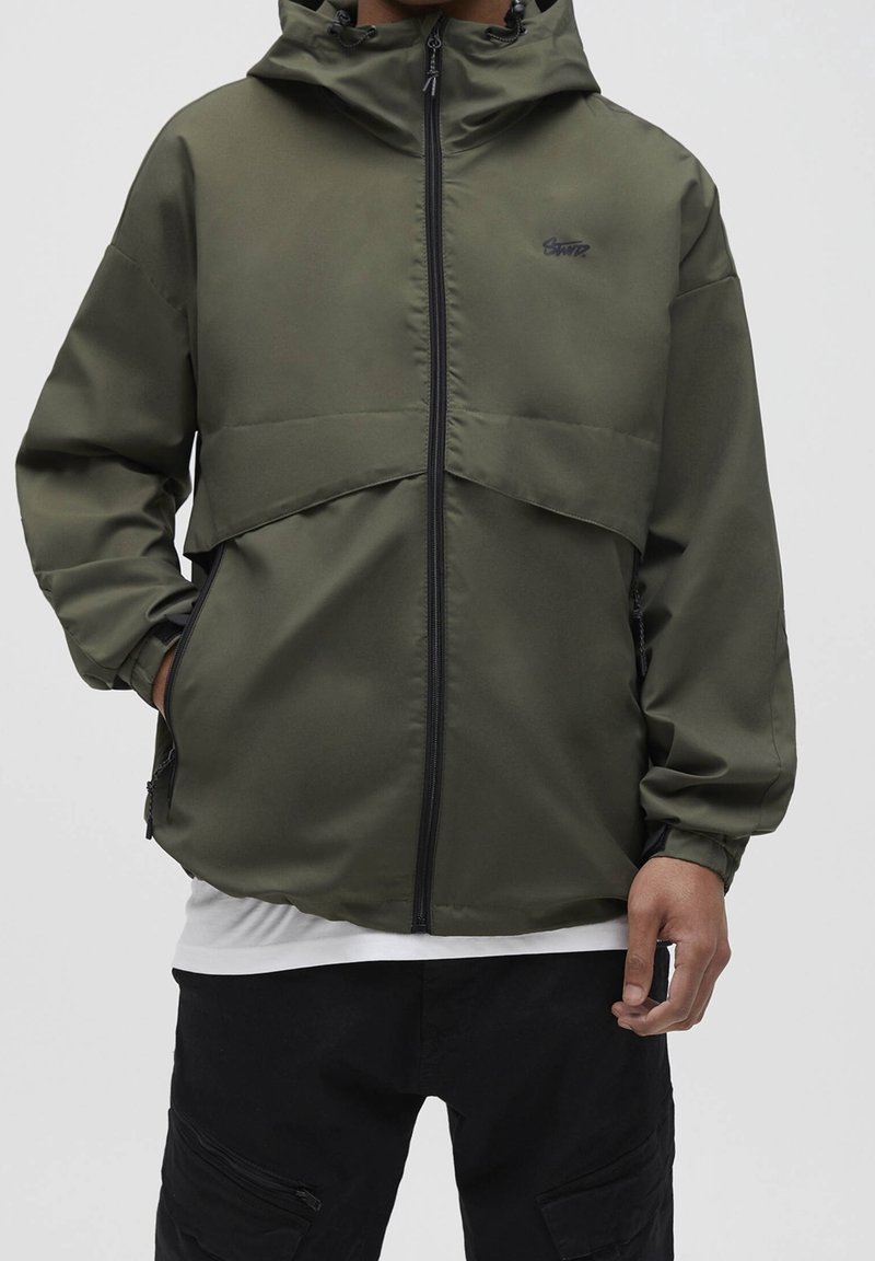 Homme portant une veste zippée à capuche vert olive avec poches latérales, pantalon cargo noir et un sous-maillot blanc visible à l'ourlet.