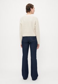 Kvinne iført en kremfarget ribbestrikket genser og mørkeblå flagrende jeans, står med ryggen til mot en enkel hvit bakgrunn.