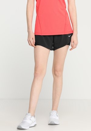 Person iført koralfarvet top uden ærmer, sorte Nike sportsshorts og hvide sneakers, som står foran en ensfarvet baggrund.