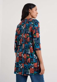 Ženska v multikolor tuniki dolgih rokavov z abstraktnimi cvetličnimi in geometrijskimi vzorci, v kombinaciji z modrimi kavbojkami, stoji bočno.