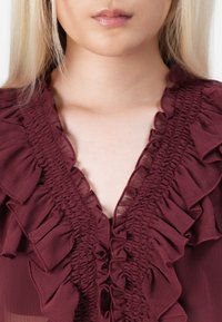 Burgunderfarget bluse med volanger langs utsnittet, teksturert stoff og dyp V-form, som fremhever et feminint design.