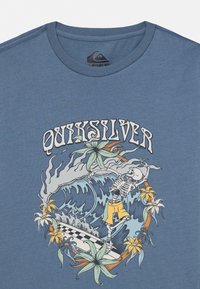 Blauwe katoenen T-shirt met een afbeelding van een skelet dat op een golf surft, omringd door florale accenten en het "Quiksilver" logo.