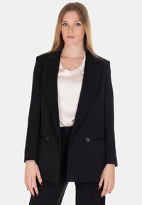 Blazer nero doppiopetto con rever a punta, due bottoni frontali e spalle strutturate. Indossato sopra una camicetta in satin bianco.
