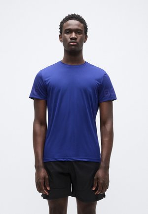 Maglietta sportiva a maniche corte blu con scollo rotondo. Tessuto liscio con dettaglio logo sulla manica sinistra. Abbinata a pantaloncini neri.