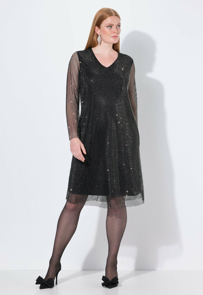 Schwarzes Kleid mit langen, durchscheinenden Netzärmeln, verziert mit Glitzerdetails. V-Ausschnitt, knielanger Saum, kombiniert mit schwarzen Schuhen mit Schleifenverzierung.