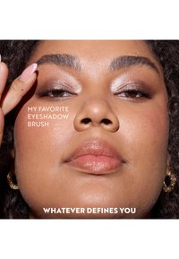 Close-up of a face with medium brown skin, wearing shimmery eyeshadow. The model has curly hair, a natural lip color, and gold hoop earrings.