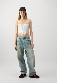 Un crop top blanc avec des coutures verticales, un jean baggy bleu clair et une casquette noire avec logo ; des baskets noires avec des accents rouges.