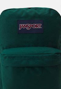 Sac à dos en tissu vert avec le logo JanSport. Il dispose d'une fermeture éclair supérieure et d'une poche avant. Texture lisse, forme arrondie et couture robuste.