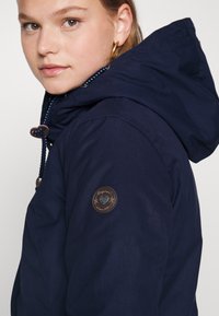 Giacca con cappuccio blu navy con finitura texture, con una patch logo marrone sulla manica e un cordino a contrasto per l'aggiustamento.