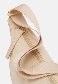 Beige látková taška se zipem a nastavitelným silným ramenním popruhem, zobrazená v detailním záběru na bílém pozadí.