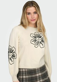 Kremfarget genser med svart blomsterbroderi på ermene, ribbestrikkede mansjetter og kant. Kombinert med et rutete skjørt med grå og beige toner.