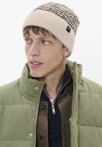 Bonnet en tricot crème avec un motif texturé en noir, porté avec une doudoune verte et un t-shirt superposé marron et bleu marine, mettant en valeur un style décontracté.