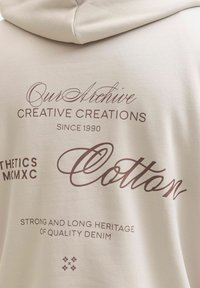 Beež puuvillane kapuutsiga pusa pruunide dekoratiivtekstidega, millel on fraasid nagu „Creative Creations“ ja „Strong Heritage of Quality Denim“.