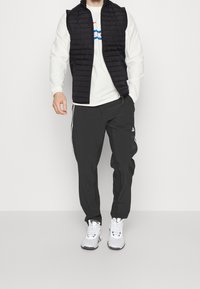 Schwarze gesteppte Weste über einem weißen Langarmshirt, kombiniert mit schwarzen Jogginghosen mit weißen Seitenstreifen und weißen Sneakers.