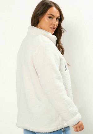 Chaqueta de forro polar blanca con un alto cuello, tejido texturizado y cordón en el dobladillo. La chaqueta tiene un material suave y esponjoso y mangas largas.