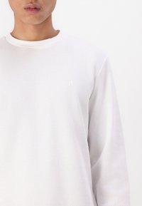 Weißes, strukturiertes Langarmshirt mit rundem Ausschnitt und kleinem, gesticktem Logo auf der Brust. Waffelstrickstoff.