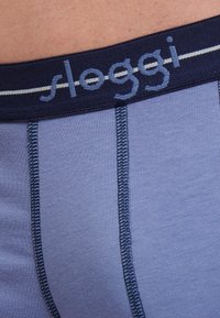 Ljusblå herrkalsonger med ett marinblått elastiskt midjeband med "sloggi"-logotyp, fina sömmar och en slät textur.