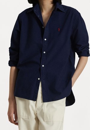 Personne portant une chemise bleu marine à boutons avec un logo rouge sur la poitrine et un pantalon couleur crème, une main dans la poche.