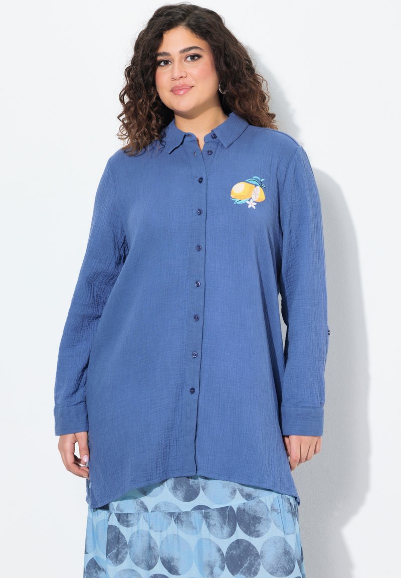 Camicia azzurra con bottoni realizzata in un tessuto testurizzato, con un ricamo di limone giallo sul petto. Maniche lunghe con polsini abbottonati.