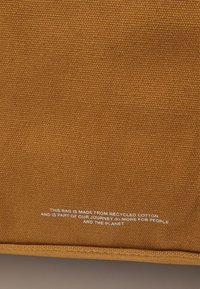 Bolsa de algodón reciclado en un cálido color mostaza con una superficie texturizada. Presenta un texto blanco que detalla los materiales sostenibles.