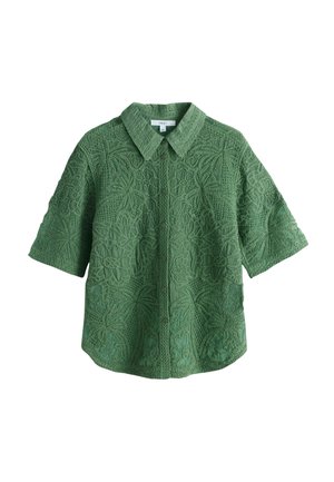 Femme marchant sur un chemin pavé portant une chemise verte texturée à manches courtes et un short assorti avec des sandales blanches à lanières en plein air.