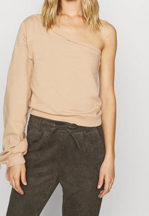 Kobieta ubrana w beżowy top na jedno ramię z długim rękawem i wiązanym mankietem na lewej ręce oraz ciemnoszare spodnie z trokiem.