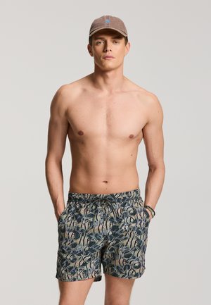 Mladý muž s hnedou čiapkou a vzorovanými plaveckými šortkami stojaci s rukami v vreckách na jednoliatom pozadí.