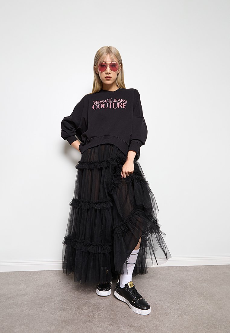Sudadera negra de gran tamaño con el texto "VERSACE JEANS COUTURE", falda de tul negro en capas y zapatillas negras con acentos blancos y tachuelas.