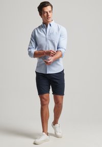 Lichtblauw button-up shirt met een borstzak, marineblauwe shorts en witte sneakers. De stof lijkt glad; mouwen opgerold tot de ellebogen.