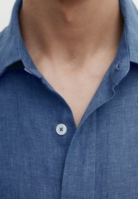 Blaues Leinenhemd mit weicher Textur, ausgestattet mit einem Kentkragen und einem einzigen Knopf am Hals. Nahaufnahme, die die Stoffdetails betont.