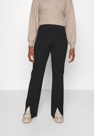 Kvinde iført beige sweater, sorte lige bukser med slidser foran over beige sneakers, står mod en ensfarvet hvid baggrund.