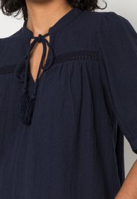 Blouse bleu marine avec un col en trou de serrure, accent en pompon, manches courtes et tissu texturé ornée de dentelle horizontale.