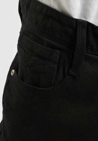 Nærbilde av svart denim bukser med midje og frontlomme, synlig søm og metallnitter, iført en lysfarget topp.