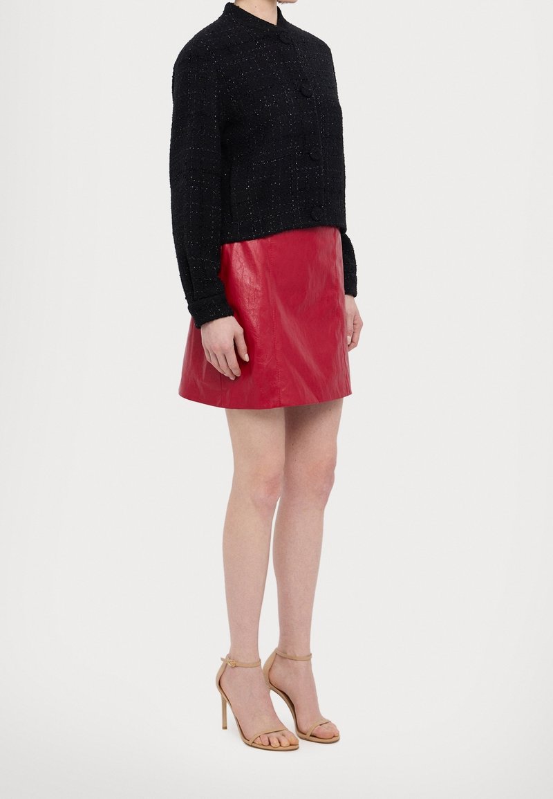 Femme portant une veste noire texturée à manches longues, une mini-jupe en cuir rouge et des sandales à talons beiges, debout devant un fond blanc.