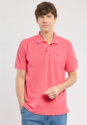 Homme portant un polo rose corail à manches courtes et un jean bleu, debout devant un fond clair uni, regardant la caméra.
