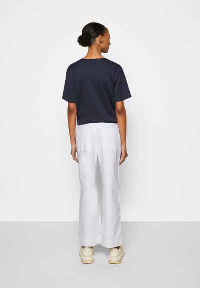 T-shirt azul-marinho de manga curta combinado com calças brancas de perna larga, com bolsos traseiros. O modelo usa ténis bege. Design minimalista.