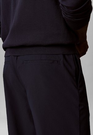 Vue de dos d'une personne portant un pantalon texturé foncé et un pull foncé assorti, la main droite reposant près de la taille.