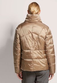 Leichte braune gepolsterte Jacke mit hohem Kragen, geometrischen Nähten und glattem, glänzendem Stoff.