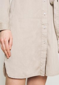Beige shirt dress in pehmeästä kankaasta, valkoisilla napilla, suora leikkaus ja sivuhalkiot liikkumisen helpottamiseksi. Yhteensopiva sileä tekstuuri.