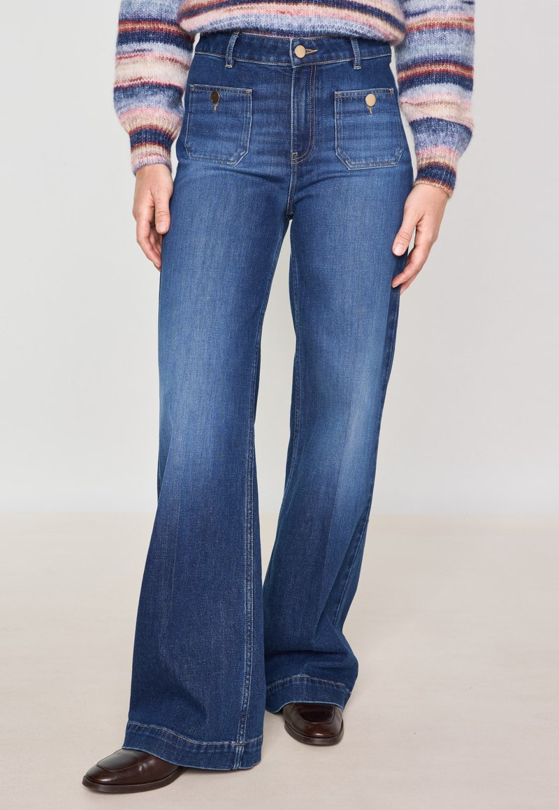 Jeans in denim a vita alta a campana di colore blu scuro, dotati di tasche frontali e dettagli in metallo dorato, con una texture liscia e leggermente scolorita.