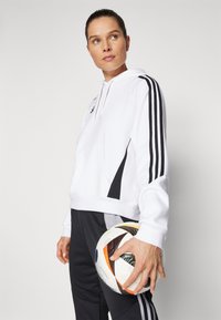 White hooded sweatshirt with black accents, featuring a front pocket and three black stripes down the sleeves. Model holds a soccer ball.