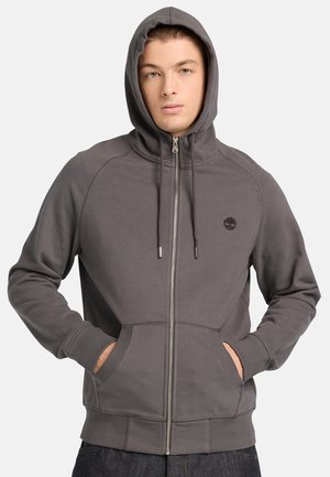 Joven con una sudadera gris con cremallera y capucha puesta, con las manos en los bolsillos delanteros, frente a un fondo liso.