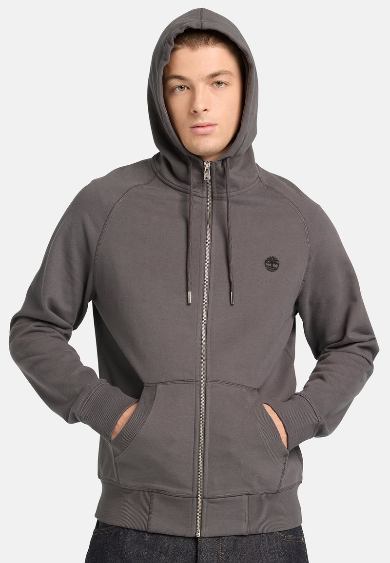 Joven con una sudadera gris con cremallera y capucha puesta, con las manos en los bolsillos delanteros, frente a un fondo liso.