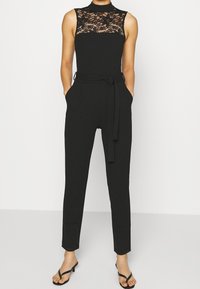 Mono negro con un alto cuello de encaje, cuerpo ajustado, piernas acampanadas, bolsillos laterales y un cinturón de lazo en la cintura. Textura suave, diseño minimalista.