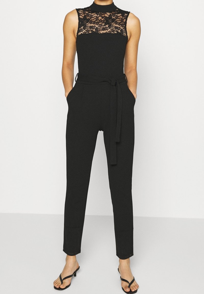 Mono negro con un alto cuello de encaje, cuerpo ajustado, piernas acampanadas, bolsillos laterales y un cinturón de lazo en la cintura. Textura suave, diseño minimalista.